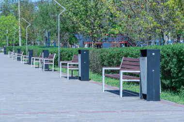 Kahverengi ahşap bankları olan şehir parkı. Kamu parkında simetri koltuğu olan bir dinlenme alanı. Yer kaldırımda oturuyor. Oturma alanı, rahatlama için rahat bir noktada ahşap banklar ve çöp kutusuna sahiptir..