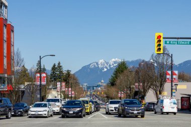 Vancouver, Kanada - 18 Mart 2023: Fotoğraf, Vancouver 'daki Kral Edward Caddesi ile Cambie Caddesi' nin kesişiminin görkemli dağların manzaralı bir zemin oluşturduğu bir görüntüsünü çeker..