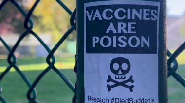 Fotoğrafta metal bir direğin üzerinde kurukafa ve çapraz kemik sembolü olan bir etiket ve kalın metin VACCINES zehirlidir
