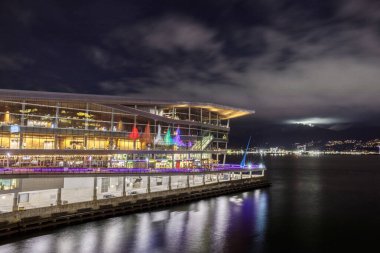 Vancouver, Kanada - 3 Ocak 2023: Vancouver Kongre Merkezi 'nin deniz manzarası ve liman sularında renkli yansımalarla aydınlatılmış yelkenler ve cam cephe, arka planda görünen dağlar