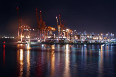 Vancouver 'ın konteynır limanının gece görüşü aydınlatmalı kargo vinçleri ve terminal ışıkları sakin liman sularında, endüstriyel deniz manzarasında altın yansımalar yaratıyor.