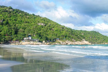 Famous Haad Rin Beach in Koh Phangan with white sandy shoreline, rolling waves, and beachfront resorts nestled against green hillside with granite boulders clipart