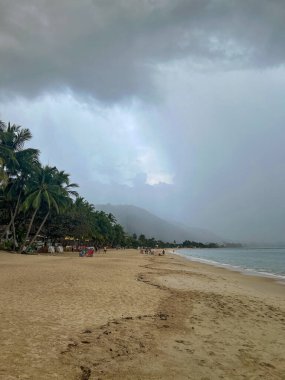 Heavy rain clouds approaching Lamai Beach with misty mountain backdrop, palm trees lining the sandy shore, and beach-goers visible along the coastline clipart