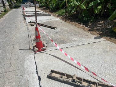 Damaged concrete road section marked with safety cone and red-white warning tape, showing cracks and exposed infrastructure requiring repair clipart