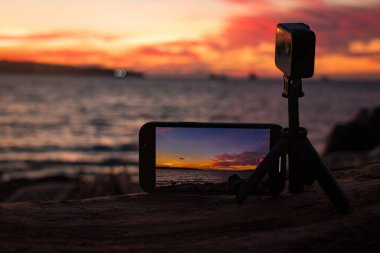 Mobil telefon ve aksiyon kameraları Vancouver sahilinde okyanus batımını görüntülüyor, dramatik turuncu ve kırmızı gökyüzü telefon ekranına yansıyor.