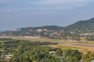 Ko Samui Havalimanı 'ndan Kao Hua Jook Pagoda' nın panoramik manzarası. Pistte, yemyeşil tropikal ormanlar, yamaçlardaki gelişmeler ve sisli mesafedeki kıyı dağları ile çevrili birçok uçak bulunuyor.
