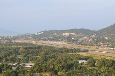 Panoramic view from Kao Hua Jook Pagoda of Ko Samui Airport with several aircraft on the runway, surrounded by lush tropical forest, hillside developments, and coastal mountains in the misty distance clipart