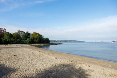 Vancouver 'daki sakin Kitsilano Sahili, sakin İngiliz Körfezi sularıyla buluşan altın kumlar, açık mavi gökyüzü altında yelkenliler, kargo gemileri ve uzak dağlarla benekli