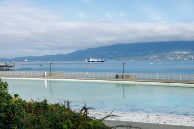 Vancouver, Kanada - 15. Sezon 2025: Kitsilano Havuzu 'nun sakin bir rıhtım, uzak dağlar ve parçalı bulutlu bir gökyüzü tarafından çerçevelenmiş yüzücülerin suyun tadını çıkardığı manzarası