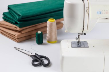 Dikiş makinesi, iplik makaraları ve keskin makasla birlikte temiz yüzeyde oturur. Yeşil ve kahverengi renklerde katlanmış kumaş, el işi ve yaratıcılığı bekliyor..