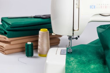 Çevredeki ipliklerin çeşitli makaralarıyla yeşil kumaşı dikebilen bir dikiş makinesi. Masanın üzerinde düzgünce katlanmış kahverengi kağıt var. Tekstil projelerine yönelik hazırlıkları gösteriyor..