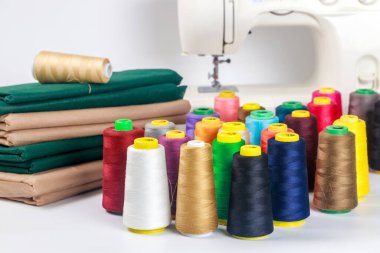 Zanaat atölyesindeki kumaş yığınlarının etrafına dizilmiş renkli iplik makaraları. Dikiş ve yaratıcılık için araçlar açıkça mevcut, projeler için hazır.