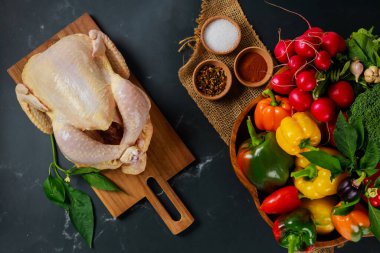 Whole chicken sits on wooden cutting board beside colorful assortment of fresh vegetables, including peppers, radishes, broccoli, ready for cooking. clipart