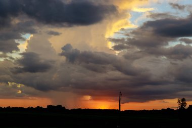 Göz kamaştırıcı günbatımı turuncu ve sarı zengin renklerle gözler önüne serilirken, koyu renk yağmur bulutları uzak mesafede bir araya gelerek, alacakaranlıkta sakin kırsal manzarayı aydınlatıyor..