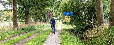 Gees, Hollanda - 11 Temmuz 2024: Hollanda 'nın Drenthe kentindeki Gees picoresque town Coevorden plasename işaretini geçen kıdemli bisikletçi.