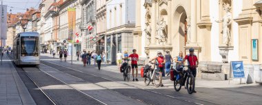 Graz, Avusturya - 6 Eylül 2024: Avusturya 'nın Stiermarken bölgesindeki Graz' da tramvayla Kutsal Kan Kilisesi 'nin önünden bisikletle geçen kıdemli gezginler