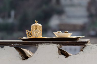 Altın ve porselenden yapılmış geleneksel bir Türk kahve seti ahşap bir tepside duruyor. Set, arka planda bulanık bir şehir manzarası olan beyaz bir duvara yerleştiriliyor..