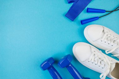 Fitness konsepti, spor. Beyaz spor ayakkabılarının, elastik bantların ve mavi halterlerin izole edilmiş mavi arkaplandaki boş alandaki en iyi görüntüsü.