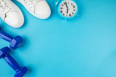 Spor aksesuarları konsepti. Beyaz alarmlı spor ayakkabıların ve mavi halterlerin izole edilmiş mavi arkaplandaki fotokopi alanının en üst görüntüsü. Antrenman zamanı..