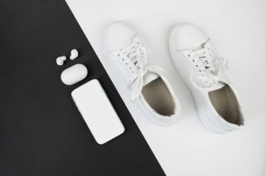 Siyah ve beyaz arka planda telefon ve kulaklıklı beyaz spor ayakkabıların fotokopi alanı ile en üstteki görünümü.