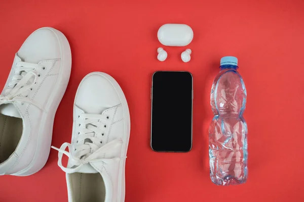 スポーツ用具 赤い背景に白いスニーカー スマートフォンやヘッドフォンのトップビュー 朝のランニング用アクセサリー — ストック写真