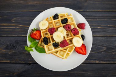 Koyu ahşap arka planda beyaz tabakta yaz meyveleri ve pudra şekerli Belçika waffleları. Kahvaltıda ya da öğle yemeğinde Belçika waffle 'ı