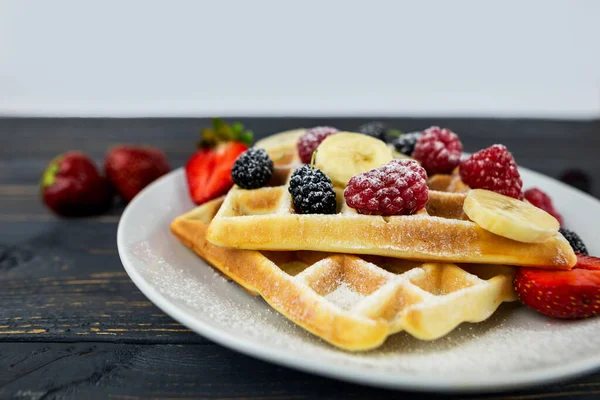 Belgian waffles with summer berries and powdered sugar in a white plate on a dark wooden background. Sweet Belgian waffles for breakfast or lunch