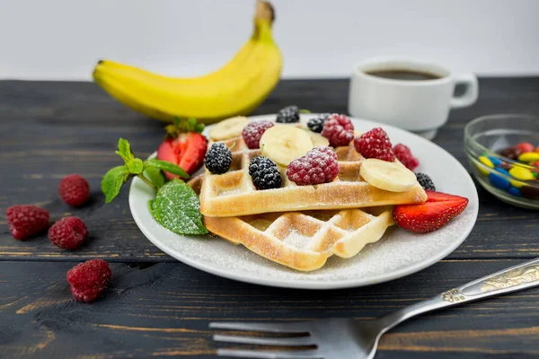 Belgian waffles with summer berries and powdered sugar in a white plate on a dark wooden background. Sweet Belgian waffles for breakfast or lunch