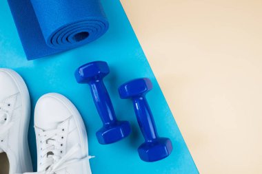 Spor aksesuarları konsepti. Beyaz spor ayakkabıları, mavi halterler, mavi spor paspası ve pastel bej arka planda diğer spor malzemelerinin bir fotoğrafı. Yukarıdan görüntüle.