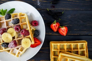 Koyu ahşap arka planda beyaz tabakta yaz meyveleri ve pudra şekerli Belçika waffleları. Kahvaltıda ya da öğle yemeğinde Belçika waffle 'ı