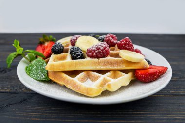 Koyu ahşap arka planda beyaz tabakta yaz meyveleri ve pudra şekerli Belçika waffleları. Kahvaltıda ya da öğle yemeğinde Belçika waffle 'ı