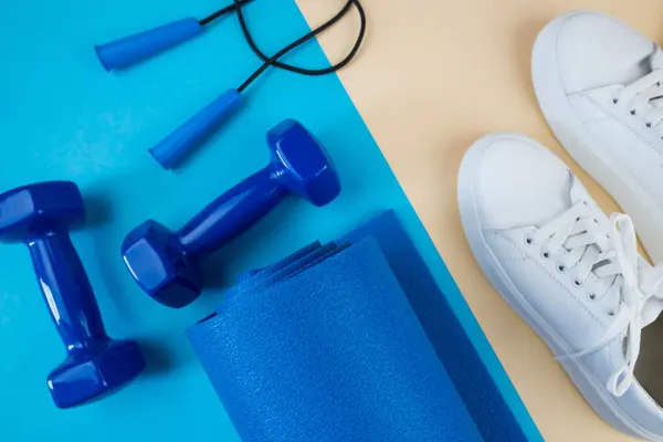 The concept of sports accessories. A photo of white sneakers, blue dumbbells, a blue exercise mat and other sports equipment against a pastel beige background. View from above.
