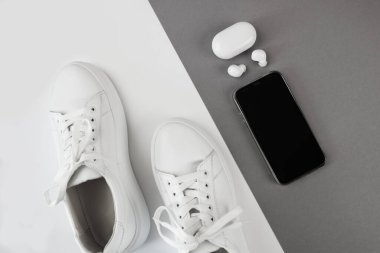 Kulaklıklı beyaz spor ayakkabılar ve gri-beyaz arka planda akıllı bir telefon. Yukarıdan görüntüle.
