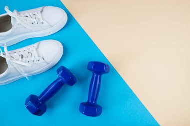 Beyaz spor ayakkabıların ve mavi halterlerin mavi-bej arkaplandaki fotokopi alanı ile en üstteki görüntüsü.