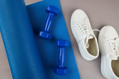 Mavi renkli yoga minderi, beyaz spor ayakkabıları, dambıllar, sağlıklı yaşam tarzı, spor ve egzersiz konsepti. Gri arkaplan.