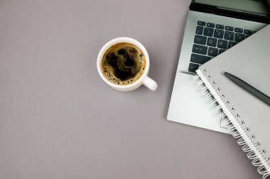 Açık dizüstü bilgisayar, bir fincan kahve, gri arka planda not defteri ve kalem. Kopyalama alanı olan üst görünüm, düz yatay