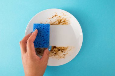 Bir kadın yemekten sonra yemek lekelerini temizlemek için mavi bir sünger ve beyaz bir tabağın üzerindeki kiri kullanır..