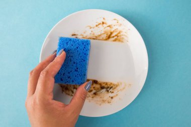 Bir kadın yemekten sonra yemek lekelerini temizlemek için mavi bir sünger ve beyaz bir tabağın üzerindeki kiri kullanır..