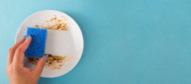 Bir kadın yemek yedikten sonra yemek lekelerini ve kiri temizlemek için mavi bir temizlik süngeri kullanır. Uzayı kopyala Metin için yer.