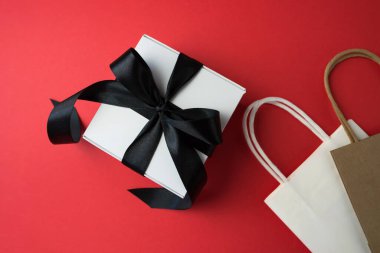 Siyah kurdeleli hediye kutusu ve kırmızı arka planda fotokopi alanı olan alışveriş torbaları. Kara Cuma indirimi.