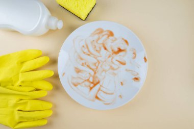 Kirli, beyaz bir tabak bulaşık deterjanı, sarı süngerler ve bej arka planda eldivenler. Üst manzara. Bulaşıkları yıka..