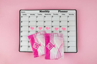 Beyaz takvim ve açık pembe arka planda bir paket hijyenik ped. Pastel renk. Yakın plan. Adet döngüsü.