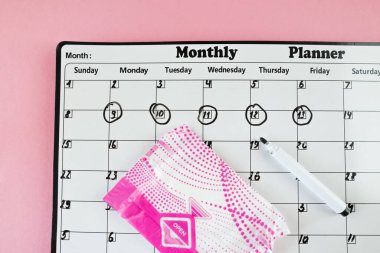 Beyaz takvim ve açık pembe arka planda bir paket hijyenik ped. Pastel renk. Yakın plan. Adet döngüsü.
