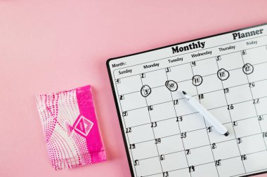 Beyaz takvim ve açık pembe arka planda bir paket hijyenik ped. Pastel renk. Yakın plan. Adet döngüsü.