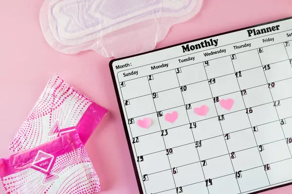Beyaz takvim ve açık pembe arka planda bir paket hijyenik ped. Pastel renk. Yakın plan. Adet döngüsü.