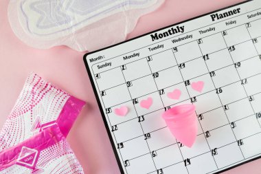 Beyaz takvim ve açık pembe arka planda bir paket hijyenik ped. Pastel renk. Yakın plan. Adet döngüsü.