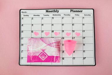Beyaz takvim ve açık pembe arka planda bir paket hijyenik ped. Pastel renk. Yakın plan. Adet döngüsü.