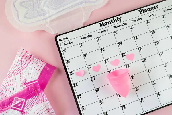 Beyaz takvim ve açık pembe arka planda bir paket hijyenik ped. Pastel renk. Yakın plan. Adet döngüsü.