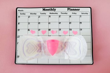 Beyaz takvim ve açık pembe arka planda bir paket hijyenik ped. Pastel renk. Yakın plan. Adet döngüsü.