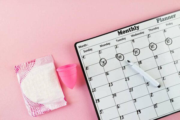 Белый календарь и пачка гигиенических прокладок на светло-розовом фоне. Цвет пастели. Крупный план. Менструальный цикл.